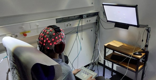 Mensch sitzt für EEG-Messung mit Elektroden auf dem Kopf in Untersuchungsraum und guckt auf Bildschirm