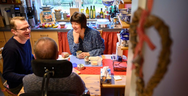 Tomas Gerlach im Rollstuhl mit Ehefrau und Betreuer lachend am Frühstückstisch