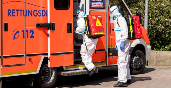 Stefane Beck und ihr Kollege, Notfallsanitäter Sebastian Götsch, steigen in den Rettungswagen. Sie tragen Schutzkleidung, die sie komplett verhüllen, und riesige rote Rucksäcke
