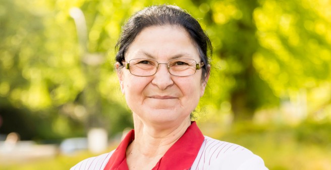 Frau Beyazgül blickt forsch in die Kamera. EIn Porträt. Schwarze, lange Haare zum Zof gebunden, rechteckige Brille. trägt die Uniform für Pflegekräfte, weiß mit zarten roten Streifen und einem roten Kragen.  sie steht im Grünen