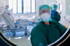 Frau Beyagül blickt durch das Bullauge der geschlossenen Tür eines sehr hellen, weißen Intensiv-Zimmers. rechts hinter ihr eine Pflegerin, links das Bett eines Patienten, dessen Bett-Rückenlehne hoch gestellt ist.