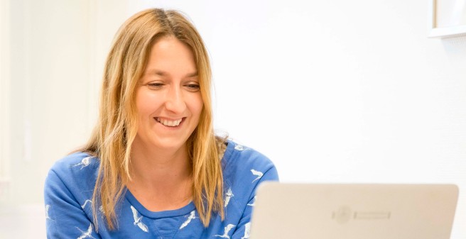 Frau Dr. Aden sitzt leicht vorgebeugt an einem kleinen Tisch und blickt lächelnd auf den Monitor des vor ihr stehenden, aufgeklappten Laptops. 