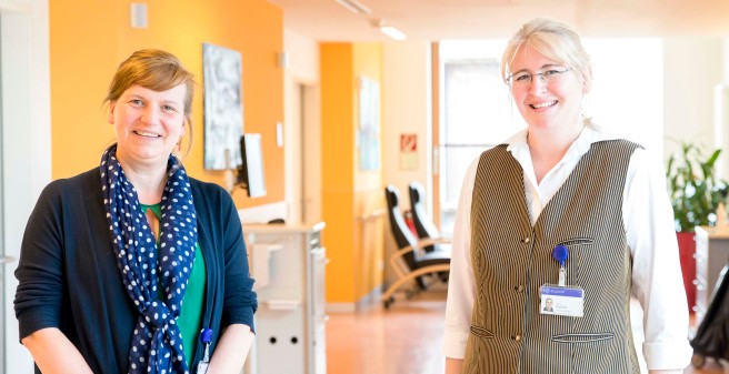 Bianca Stuchlik und ihre Kollegin Yvonne Habermann blicken lächelnd ein die Kamera. Frau Stuchlik trägt eine blaue Strickjacke und eine blauen Schal mit großen Tupfen, Frau Habermann rechts daneben, die ARbeitskleidung der Gastro Mitarbeiter: weißes Hemd, getreifte Weste über einer pinkfarbenen Schürze