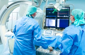 Dr. Marco Remmel im Katheter Labor bei der Arbeit, er und sein Kollege in blauer Schutzkleidung, sie blicken auf einen Monitor