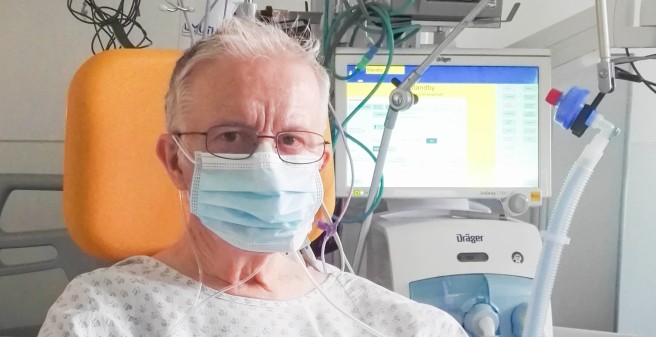 Peter Seifert auf der Intensivstation. Er trägt eine Mund-NAsen-Schutzmaske. Aus dunken Augen blickt Seifert erschöpft in die Kamera. Die Hände sind liegen flach auf der Ablage des Sitzes. Sie sind bandagiert und die Finger mit technischen Geräten verknüpft 