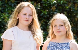 Lena, in einem weißen Shirt und Sophia, in einem blauen Kleid, stehen nebeneinander und blicken in die Kamera. Beide haben lange blonde Haare.