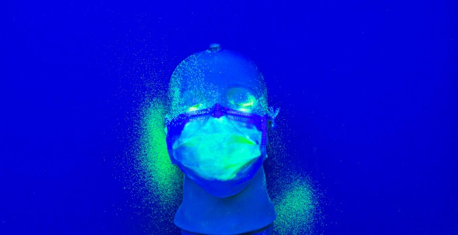 Auf einem tiefblauen Untergrund liegt ein Kunststoff-Kopf mit einer Mund-Nasenschutz-Maske. Auf der Maske, sowie links und rechts auf der Unterlage sind hellgrüne Punkte verstreut