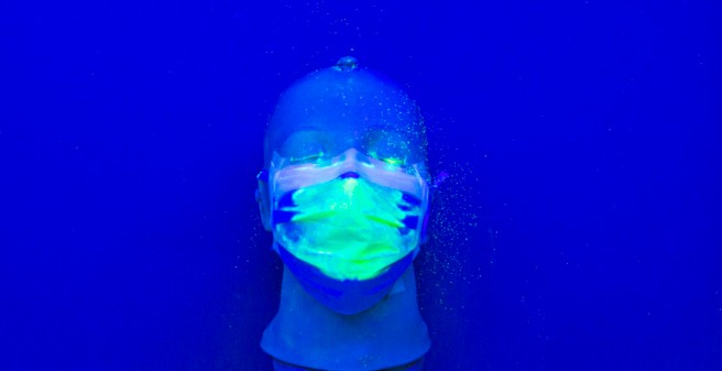 Auf einem tiefblauen Untergrund liegt ein Kunststoff-Kopf mit einer Mund-Nasenschutz-Maske. Auf der Maske sind hellgrüne Punkte