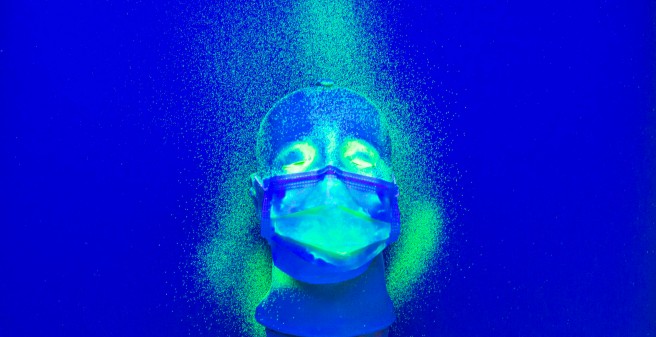 Auf einem tiefblauen Untergrund liegt ein Kunststoff-Kopf mit einer Mund-Nasenschutz-Maske. Auf der Maske, dem oberen Teil des Gesichts und Keilförmig links und oben sind hellgrüne Punkte