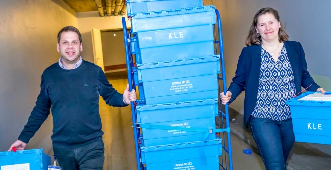 David Bielang, Laura Weinhold schieben einen Rollwagen durch einen Liefergang des UKE, der Wagen ist voll beladen mit blauen Kisten