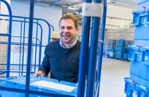 Daniel Bielang, in gemustertem Hemd und blauem Pullover, schaut lächelnd in die Kamera. Er stellt gerade eine blaue Kiste in einen Rollconteiner. Rechts von ihm ein hoher Stapel weiterer Kisten. 