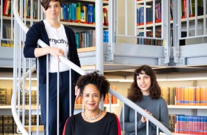 (v.l.): Dr. Christine Dahlke, Prof. Dr. Marylyn Addo, im Vordergrund, und Dr. Anahita Fathi stehen au feiner Wendeltreppe in einer Bibliothek und blicken lächelnd in die Kamera