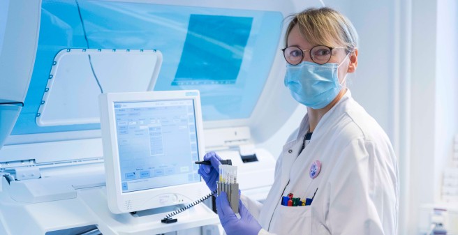 Eine Mitarbeiterin in Schutzkleidungs steht vor einem sehr großen technischen Gerät und gibt Informationen zu Probenröhrchen ein, die sie in der Hand hält
