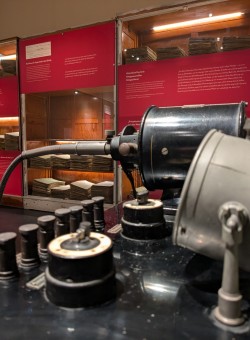 Schwarzes Pantostat-Gerät vor einer beleuchteten, rot bekleten Vitrine mit gestapelten Krankenakten