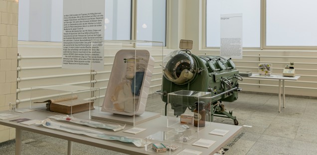  Eine Eiserne Lunge aus den 1950er Jahren zwischen Ausstellungstischen, im Vordergrund ein modernes ECMO-KIt aus der Intensivmedizin