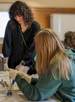 Eine stehende MItarbeiterin des Museums zeigt sitzenden Schüler:innen Objekte auf einem Tisch