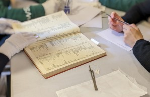 Auf einem Tisch liegt ein historischer Katalog und verschiedene Dokumente, darum die Hände von Schüler:innen
