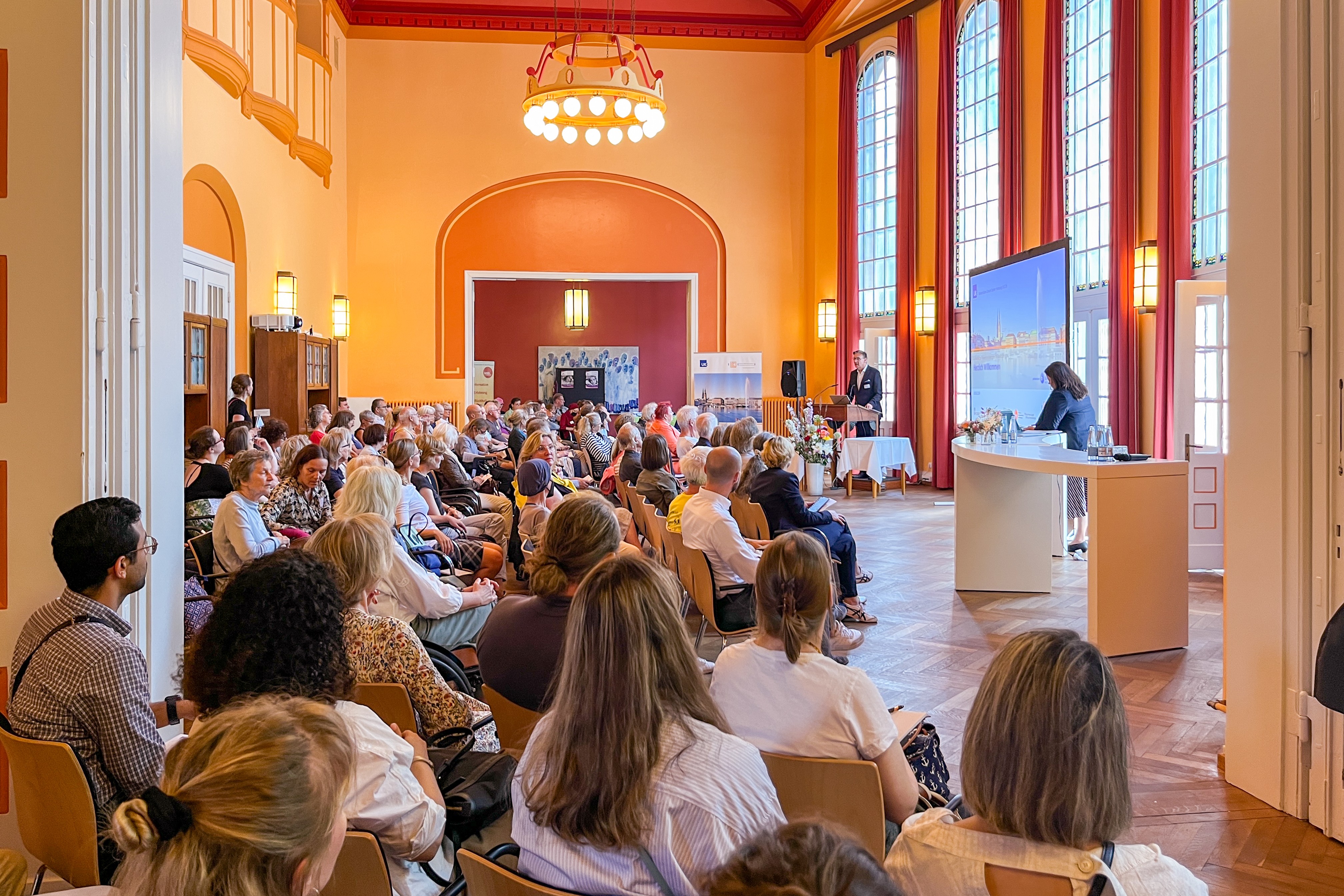 Großer Veranstaltungssaal im historischen Gebäude mit vielen Besuchern und einem Vortragenden auf der Bühne.