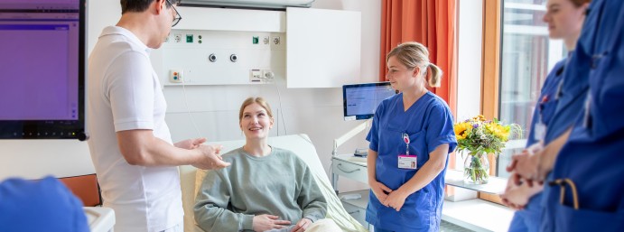 Ein Arzt im Gespräch mit einer Patientin, begleitet von Stationspflegepersonal im Rahmen der morgendlichen Visite